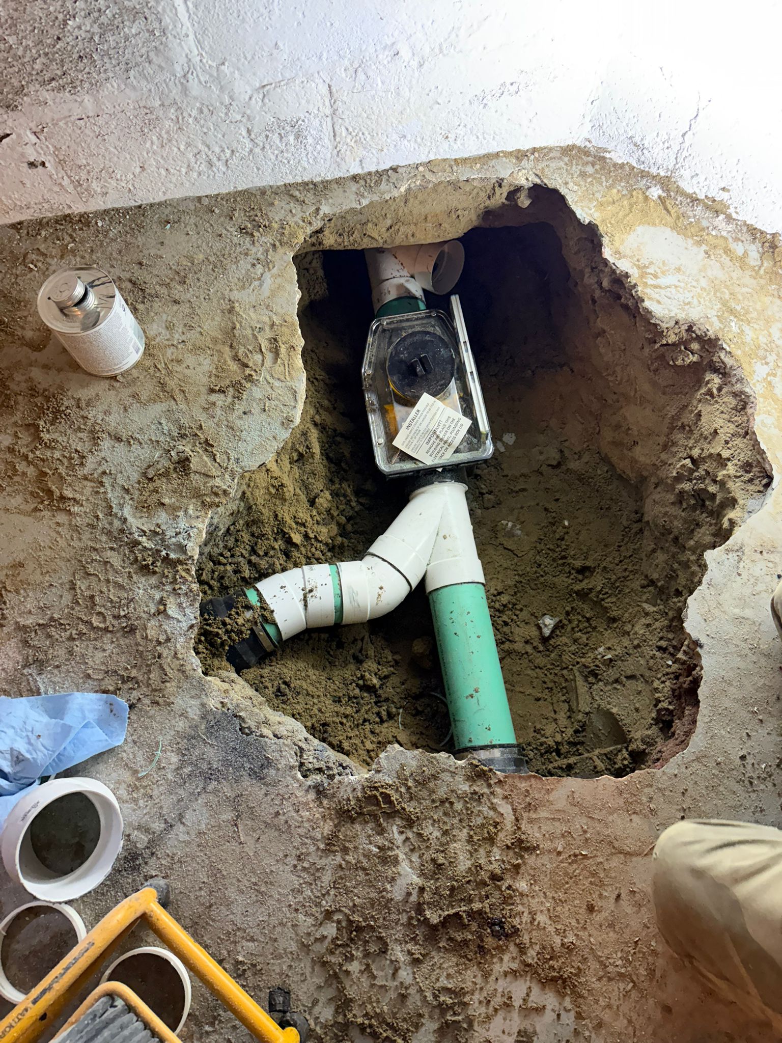 Clear-lid mainline backwater valve installed in a hole cut through a basement concrete floor, connected to green PVC drain pipe with a cleanout branch