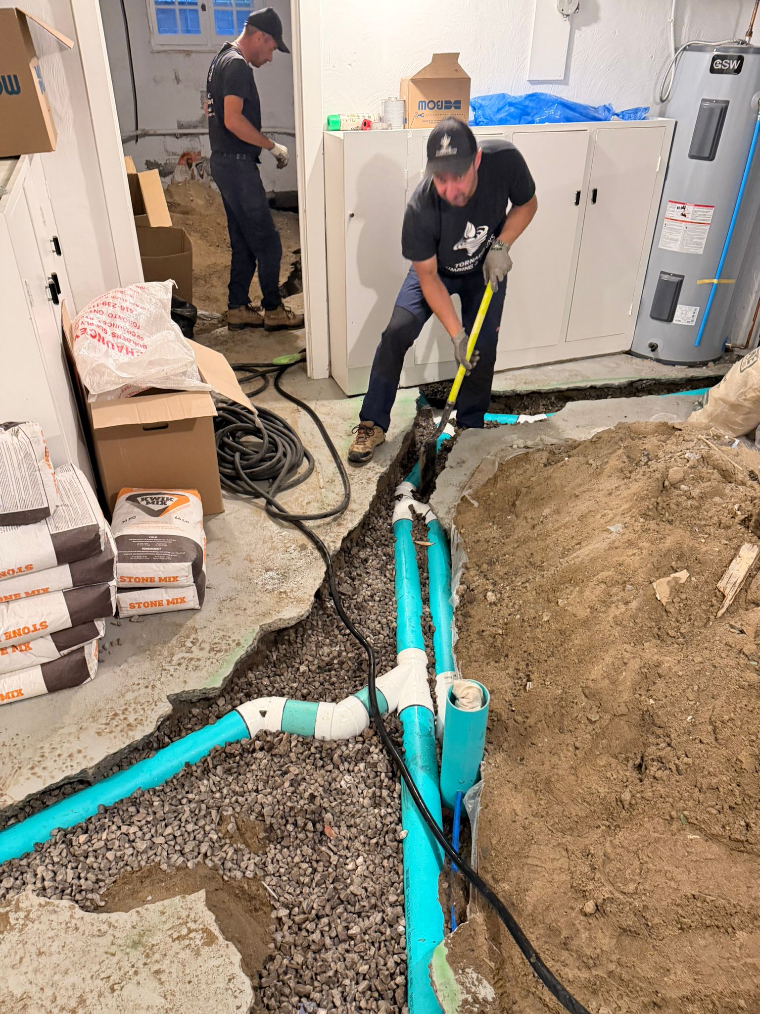 Two Tornado Plumbing technicians in a basement with the concrete floor cut open, new green PVC drain lines and wye fittings laid in a gravel trench beside a water heater