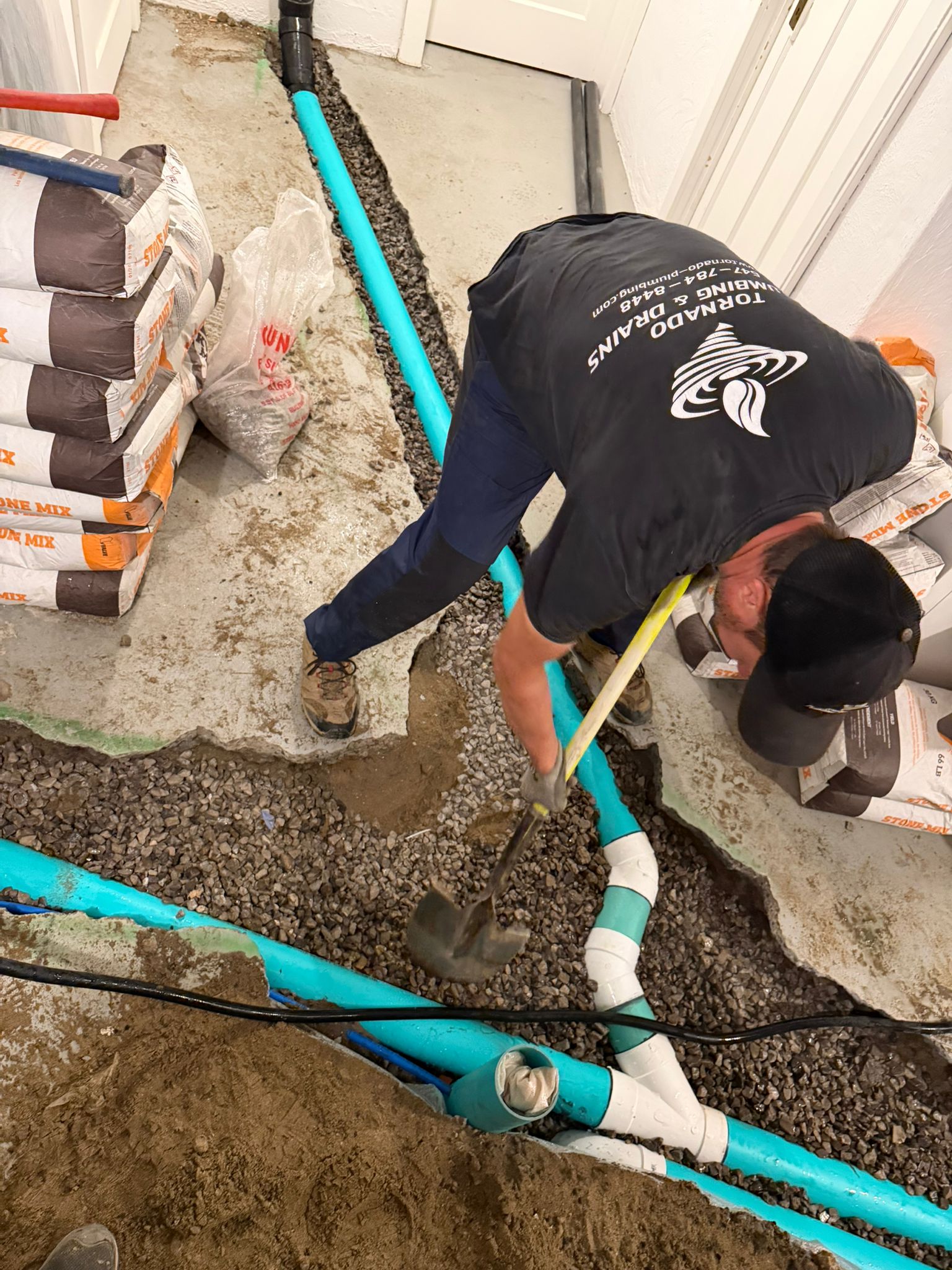 Technician shovelling gravel around new green PVC drain pipe and fittings in an open basement floor trench, stone mix bags stacked beside him