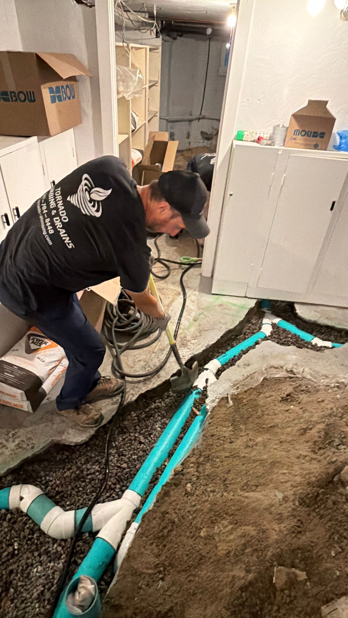 Technician in a Tornado Plumbing and Drains shirt working in a cut basement floor trench with new green PVC drain pipe running along the wall toward a stub-up