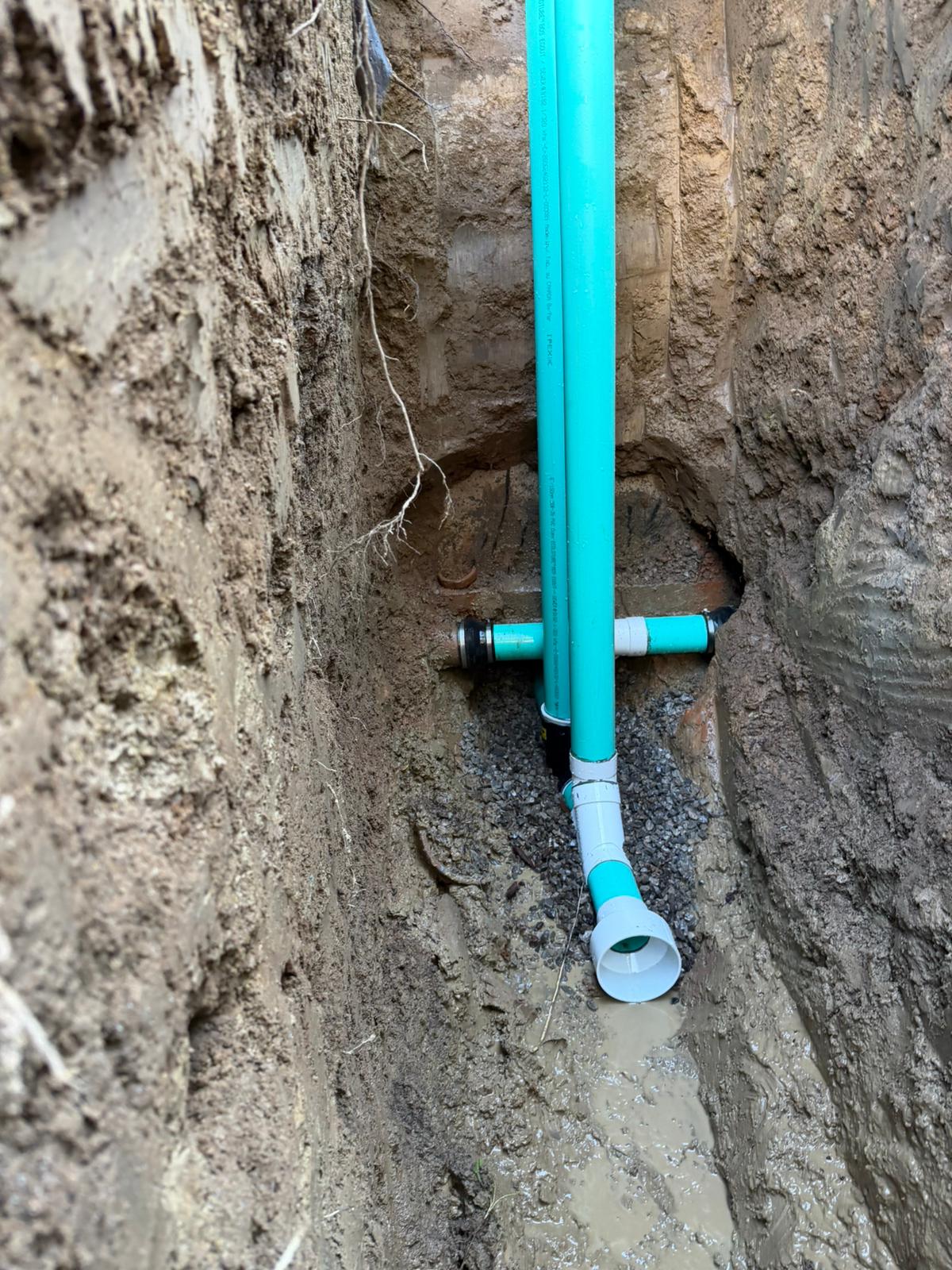 Two green PVC sewer pipes in a narrow trench joined with rubber couplings to the existing line, with a white cleanout stub pointing up and gravel underneath