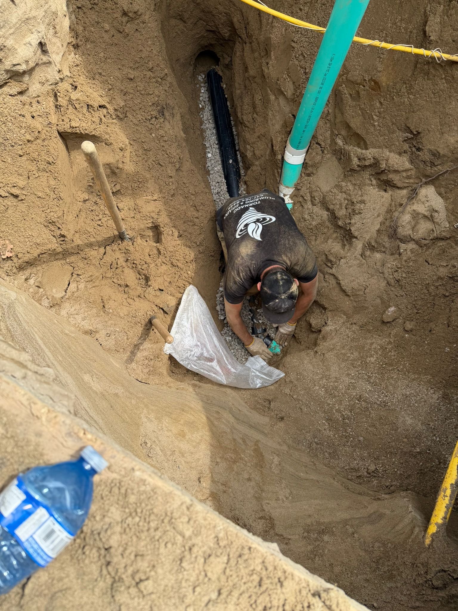 Tornado Plumbing technician kneeling in a deep sewer excavation, setting new sewer pipe on a gravel bed with a green PVC riser beside him