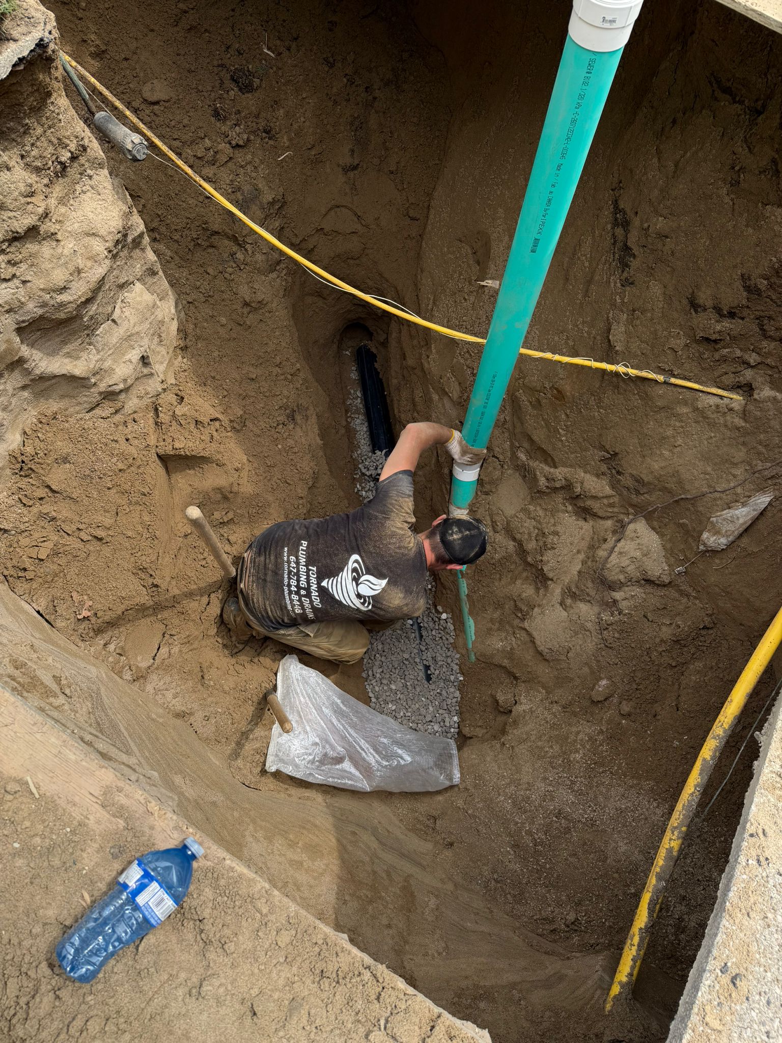 Technician in a Tornado Plumbing and Drains shirt connecting a green PVC cleanout riser to the new sewer line at the bottom of a deep trench