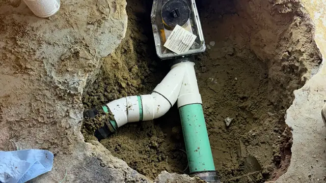 Clear-lid mainline backwater valve installed in a hole cut through a basement concrete floor, connected to green PVC drain pipe with a cleanout branch