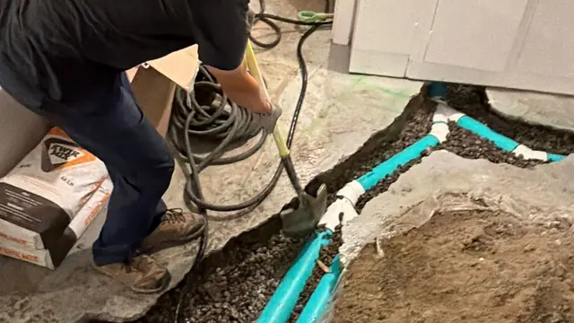 Technician in a Tornado Plumbing and Drains shirt working in a cut basement floor trench with new green PVC drain pipe running along the wall toward a stub-up
