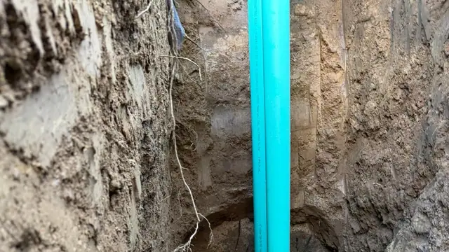 Two green PVC sewer pipes in a narrow trench joined with rubber couplings to the existing line, with a white cleanout stub pointing up and gravel underneath